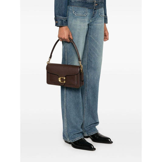 Person holding Coach Tabby 26 leather shoulder bag in earth brown pebbled leather with gold logo plaque and detachable straps.