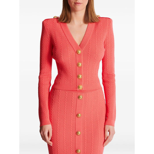 Woman wearing a coral pink Balmain Long-sleeved twisted knit cardigan with logo-embossed buttons and epaulettes, front view
