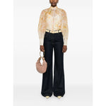 Woman wearing Zimmermann Crush Shirt in light yellow, featuring floral print, puff sleeves, crystal buttons, styled with dark wide-leg pants.